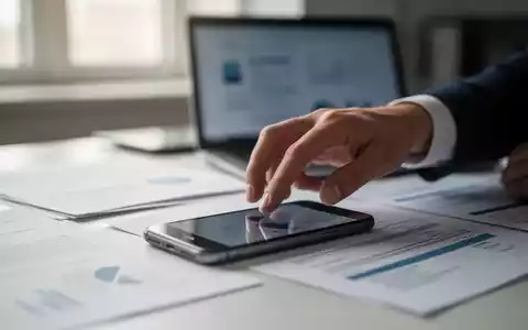 Eine Hand greift nach einem Smartphone auf einem Schreibtisch, umgeben von verschwommenen Dokumenten und einem Laptop. Fokus auf Ablenkung. - Foto: über boerse-global.de