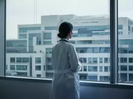 Eine Ärztin im weißen Kittel steht vor einem Fenster und blickt auf ein modernes Krankenhausgebäude. - Foto: über boerse-global.de
