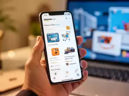 Eine Hand hält ein Smartphone mit der Amazon Shopping App, im Hintergrund verschwommene Elemente des Online-Handels. - Foto: über boerse-global.de