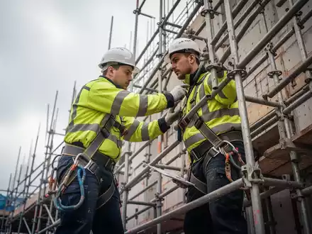 Ein Bauarbeiter in Schutzkleidung prüft ein Baugerüst auf einer Baustelle. Fokus liegt auf Sicherheit und Unfallverhütung. - Foto: über boerse-global.de