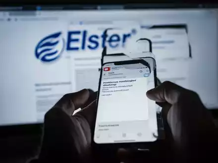 Eine Hand hält ein Smartphone mit einer gefälschten Elster-Phishing-E-Mail, im Hintergrund ein unscharfer Computerbildschirm. - Foto: über boerse-global.de