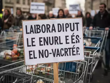 Nahaufnahme eines Protestschilds mit unscharfen Einkaufswagen und Personen im Hintergrund, das einen Arbeitskampf darstellt. - Foto: über boerse-global.de