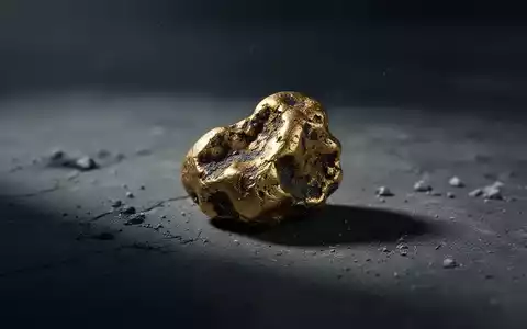 Ein einzelner, verwitterter Goldklumpen liegt auf einer dunklen, texturierten Oberfläche, teilweise im Schatten. - Foto: über boerse-global.de