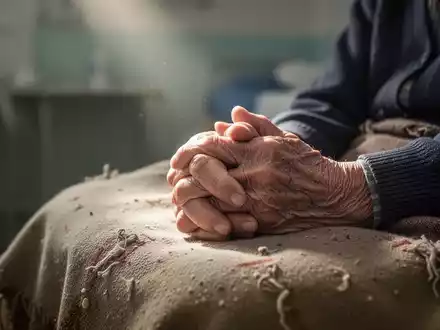 Nahaufnahme der Hände einer älteren Person, die auf einer Decke ruhen, mit einem weichgezeichneten Hintergrund, der eine Pflegeeinrichtung andeutet. - Foto: über boerse-global.de