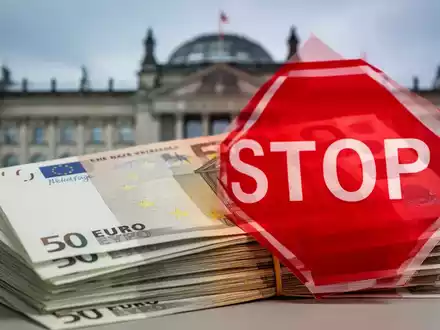 Ein Stapel Euro-Banknoten, teilweise verdeckt von einem roten Stoppschild, mit einem unscharfen Regierungsgebäude im Hintergrund. - Foto: über boerse-global.de