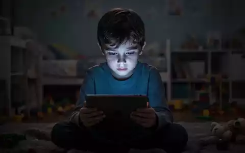 Ein Kind sitzt allein im Dunkeln, nur vom Tablet-Bildschirm beleuchtet, mit nachdenklichem Ausdruck. - Foto: über boerse-global.de