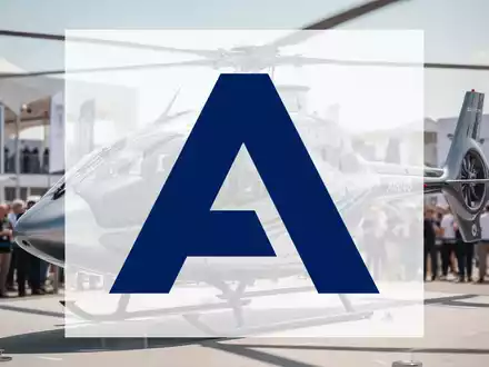 Ein moderner Airbus U145 Hubschrauber auf einer Luftfahrtmesse, mit verschwommenen Menschen und Ausstellungsgebäuden im Hintergrund. - Foto: über boerse-global.de