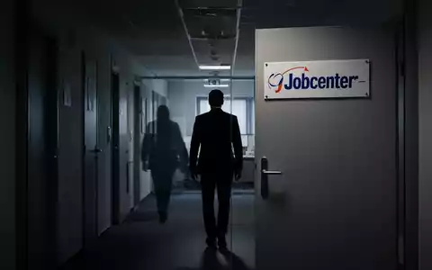Eine Silhouette geht einen dunklen Büroflur entlang, weg von einer geschlossenen Tür mit einem „Jobcenter“-Schild. - Foto: über boerse-global.de