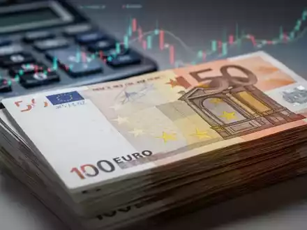 Nahaufnahme eines Stapels Euro-Banknoten, im Hintergrund verschwommen ein Taschenrechner oder Finanzdiagramm, das Löhne und Daten symbolisiert. - Foto: über boerse-global.de