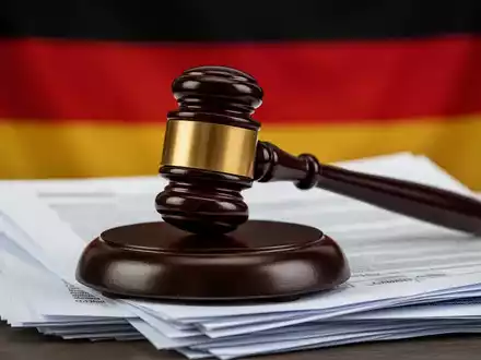 Ein hölzerner Hammer liegt auf einem Stapel Dokumente, mit der deutschen Flagge unscharf im Hintergrund, symbolisiert Gesetzgebung und Finanzen. - Foto: über boerse-global.de