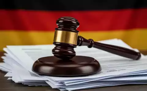 Ein hölzerner Hammer liegt auf einem Stapel Dokumente, mit der deutschen Flagge unscharf im Hintergrund, symbolisiert Gesetzgebung und Finanzen. - Foto: über boerse-global.de