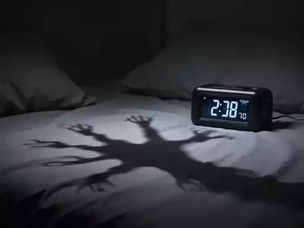 Ein dunkles Schlafzimmer mit einer digitalen Uhr, die frühe Morgenstunden anzeigt, und einem Schatten auf dem Bett. - Foto: über boerse-global.de