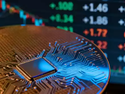 Nahaufnahme einer Siliziumwafer mit komplexen Schaltkreismustern, die blaues Licht reflektieren. Im Hintergrund ein unscharfer Börsenticker. - Foto: über boerse-global.de