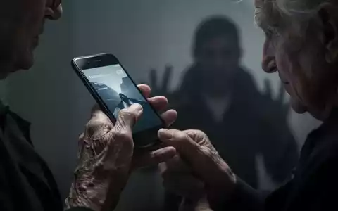 Eine ältere Hand hält ein Smartphone, im Hintergrund eine verschwommene, schattenhafte Figur, die Bedrohung und Verletzlichkeit symbolisiert. - Foto: über boerse-global.de
