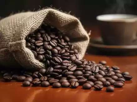 Dunkel geröstete Kaffeebohnen, die aus einem Jutesack auf eine Holzoberfläche fallen, mit einem unscharfen Kaffeebecher im Hintergrund. - Foto: über boerse-global.de