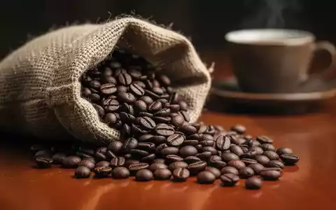 Dunkel geröstete Kaffeebohnen, die aus einem Jutesack auf eine Holzoberfläche fallen, mit einem unscharfen Kaffeebecher im Hintergrund. - Foto: über boerse-global.de