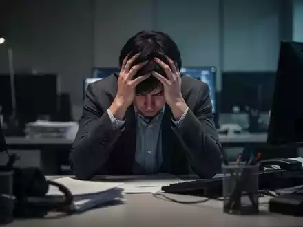 Eine Person sitzt mit dem Kopf in den Händen an einem Schreibtisch, umgeben von verschwommener Büroausstattung, was Stress und Burnout vermittelt. - Foto: über boerse-global.de