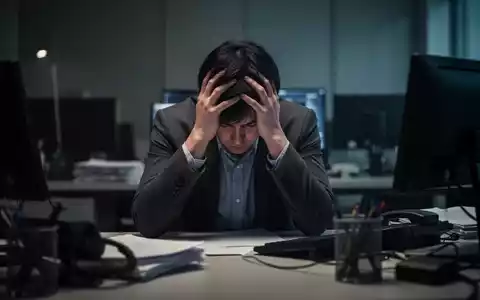 Eine Person sitzt mit dem Kopf in den Händen an einem Schreibtisch, umgeben von verschwommener Büroausstattung, was Stress und Burnout vermittelt. - Foto: über boerse-global.de