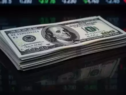 Ein Stapel neuer 100-Dollar-Scheine mit einer subtilen Spiegelung eines Börsentickers auf einer dunklen Oberfläche. - Foto: über boerse-global.de