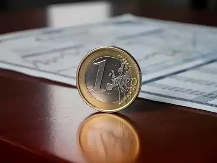 Eine einzelne glänzende Euro-Münze steht auf einem dunklen Holztisch, im Hintergrund unscharf ein Finanzbericht. - Foto: über boerse-global.de
