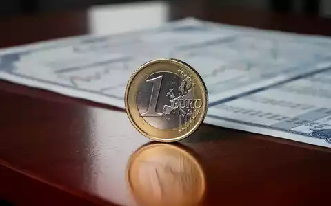 Eine einzelne glänzende Euro-Münze steht auf einem dunklen Holztisch, im Hintergrund unscharf ein Finanzbericht. - Foto: über boerse-global.de