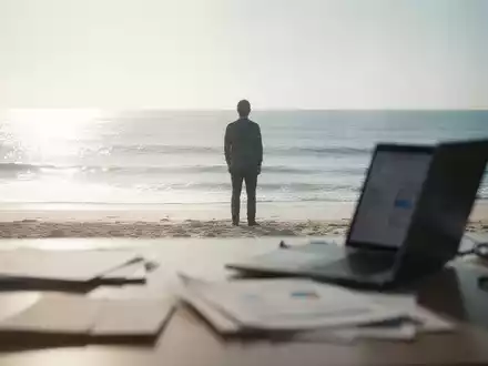 Eine Person steht am Strand und blickt aufs Meer, während im Vordergrund ein unscharfer Schreibtisch mit Laptop zu sehen ist. - Foto: über boerse-global.de