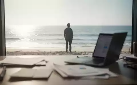Eine Person steht am Strand und blickt aufs Meer, während im Vordergrund ein unscharfer Schreibtisch mit Laptop zu sehen ist. - Foto: über boerse-global.de