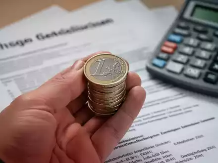 Eine Nahaufnahme einer Hand, die Euro-Münzen hält, mit verschwommenen deutschen Rechtsdokumenten und einem Taschenrechner im Hintergrund. - Foto: über boerse-global.de