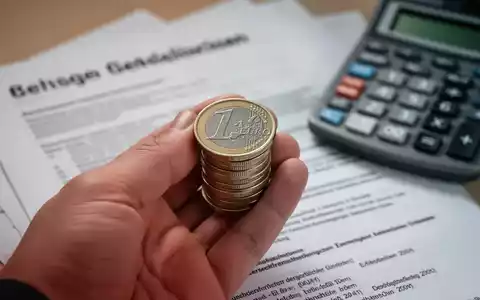 Eine Nahaufnahme einer Hand, die Euro-Münzen hält, mit verschwommenen deutschen Rechtsdokumenten und einem Taschenrechner im Hintergrund. - Foto: über boerse-global.de