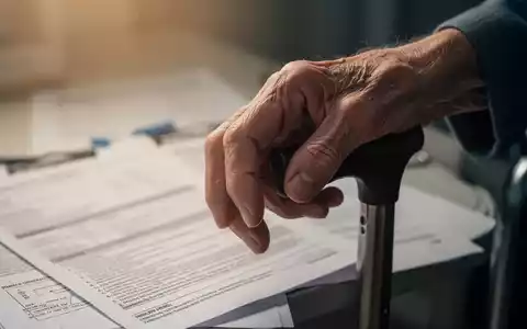 Eine ältere Hand greift vorsichtig nach einem Gehstock, im Hintergrund verschwommen medizinische Dokumente. - Foto: über boerse-global.de