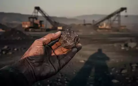 Eine verwitterte Hand hält ein kleines Stück rohes Erz vor einer unscharfen, trostlosen Bergbaulandschaft bei Sonnenuntergang. - Foto: über boerse-global.de