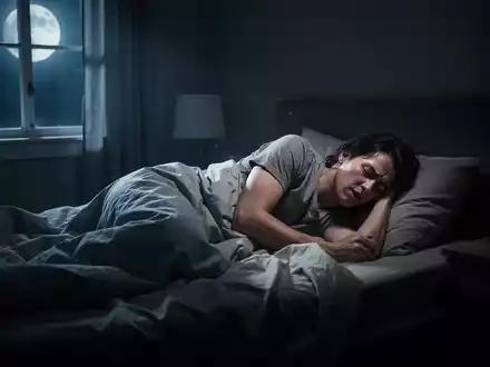 Eine Person wälzt sich unruhig in einem dunklen Schlafzimmer im Bett, beleuchtet von Mondlicht, das durch ein Fenster fällt. - Foto: über boerse-global.de