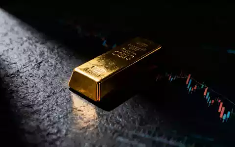 Ein goldener Barren auf dunklem Untergrund, teilweise von Schatten verdeckt, mit verschwommenen Diagrammen im Hintergrund. - Foto: über boerse-global.de