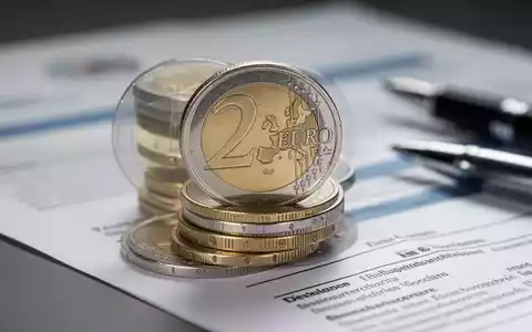 Ein Stapel deutscher Euro-Münzen mit unscharfem Hintergrund, der Dokumente und einen Stift zeigt, symbolisiert Rentendebatte. - Foto: über boerse-global.de