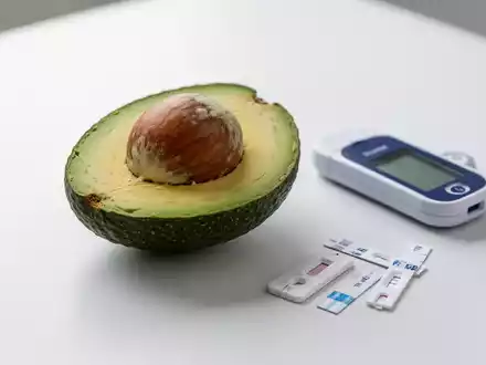 Eine halbierte Avocado mit Kern auf weißer Oberfläche, daneben ein Blutzuckermessgerät und Teststreifen. - Foto: über boerse-global.de
