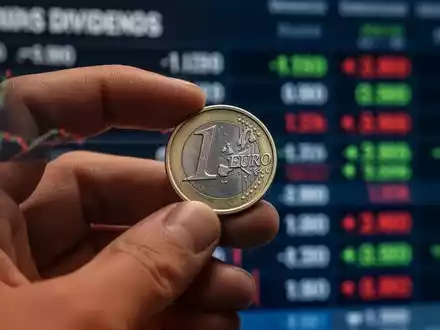 Nahaufnahme einer Hand, die eine Euromünze hält, mit verschwommenem Börsenticker im Hintergrund, der Dividenden symbolisiert. - Foto: über boerse-global.de