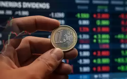 Nahaufnahme einer Hand, die eine Euromünze hält, mit verschwommenem Börsenticker im Hintergrund, der Dividenden symbolisiert. - Foto: über boerse-global.de