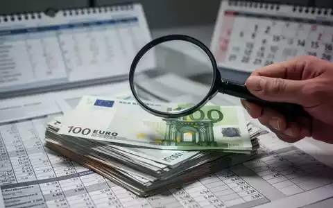 Eine Hand hält eine Lupe über Euro-Banknoten, im Hintergrund verschwommen eine Finanzübersicht und ein Kalender. - Foto: über boerse-global.de