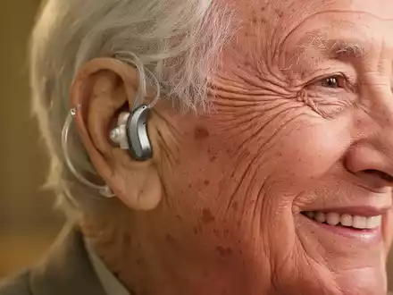 Ein älteres Ohr mit einem modernen, diskreten Hörgerät. Die Person lächelt leicht, was auf verbessertes Hören und Wohlbefinden hindeutet. - Foto: über boerse-global.de