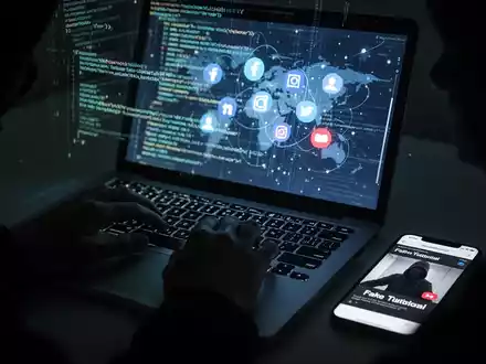 Hände tippen auf einer Laptop-Tastatur, der Bildschirm zeigt Code und Social-Media-Icons. Ein Smartphone daneben spielt ein gefälschtes Tutorial ab. Symbolisiert Cyber-Betrug und Malware-Verbreitung. - Foto: über boerse-global.de