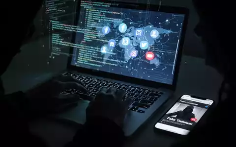 Hände tippen auf einer Laptop-Tastatur, der Bildschirm zeigt Code und Social-Media-Icons. Ein Smartphone daneben spielt ein gefälschtes Tutorial ab. Symbolisiert Cyber-Betrug und Malware-Verbreitung. - Foto: über boerse-global.de