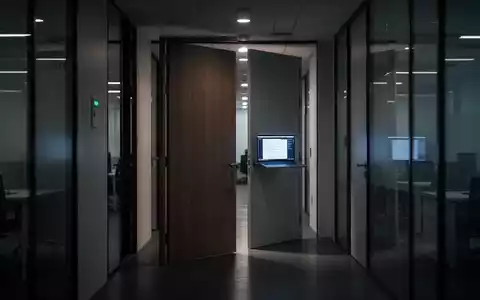 Ein leerer, modern beleuchteter Büroflur mit einer leicht geöffneten Bürotür, die einen Laptop-Bildschirm zeigt, symbolisiert Homeoffice-Verstöße. - Foto: über boerse-global.de