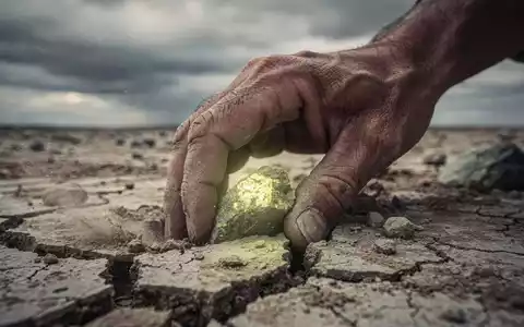 Eine einsame, verwitterte Hand siebt durch trockene Erde, die ein kleines, leuchtendes Uranerz-Nugget freilegt. - Foto: über boerse-global.de