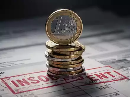 Ein unsicher gestapelter Haufen deutscher Euro-Münzen, im Hintergrund verschwommene Finanzdokumente mit einem roten 'insolvent'-Stempel. - Foto: über boerse-global.de