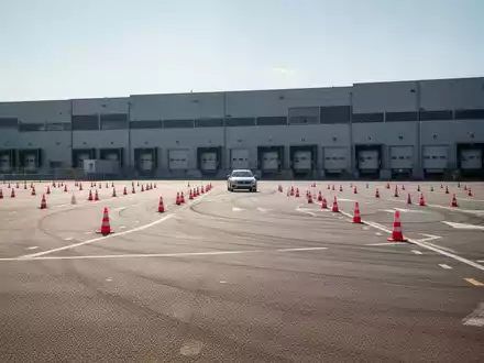 Ein leeres Asphalt-Trainingsgelände mit orangefarbenen Pylonen vor einem Logistikzentrum, bereit für ein Fahrsicherheitstraining. - Foto: über boerse-global.de