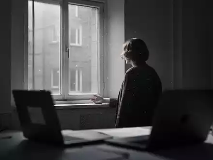Eine einsame Person blickt aus dem Fenster in einem spärlich beleuchteten Homeoffice, das soziale Isolation symbolisiert. - Foto: über boerse-global.de