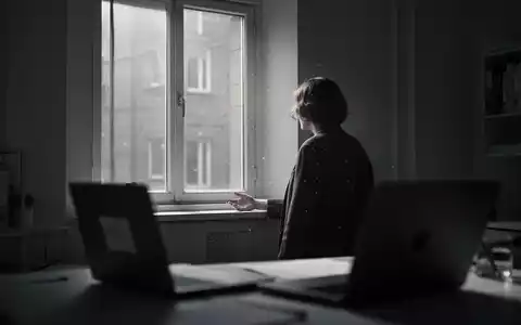 Eine einsame Person blickt aus dem Fenster in einem spärlich beleuchteten Homeoffice, das soziale Isolation symbolisiert. - Foto: über boerse-global.de
