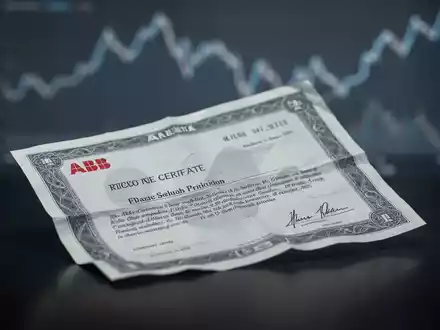 Nahaufnahme einer leicht zerknitterten ABB-Aktienurkunde auf dunklem, reflektierendem Untergrund mit fallender Trendlinie im Hintergrund. - Foto: über boerse-global.de