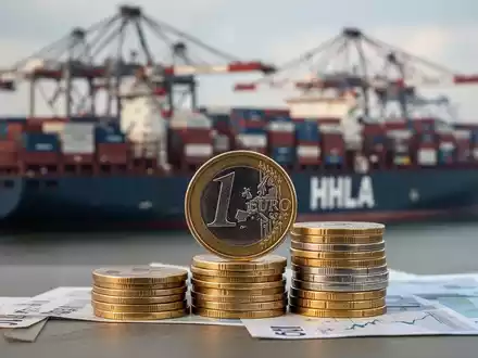 Ein Nahaufnahme von Euro-Münzen im Vordergrund, unscharf im Hintergrund ein HHLA-Containerterminal in Hamburg, bei Sonnenuntergang beleuchtet. - Foto: über boerse-global.de