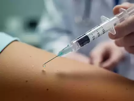 Nahaufnahme einer medizinischen Spritze mit einer Flüssigkeitstropfen an der Nadelspitze, im Hintergrund unscharf ein Arm. - Foto: über boerse-global.de